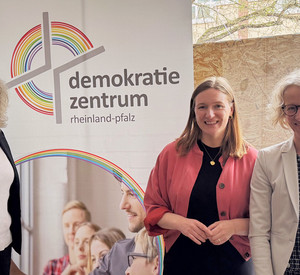Petra Fliedner, vom Demokratiezentrum, Ministerin für Familie, Frauen, Kultur und Integration, Katharina Binz, und die Präsidentin des Landesamtes für Soziales, Jugend und Versorgung, Heike Gorißen-Syrbe, vor einem Roll-Up des Demokratiezentrum Rheinland-Pfalz