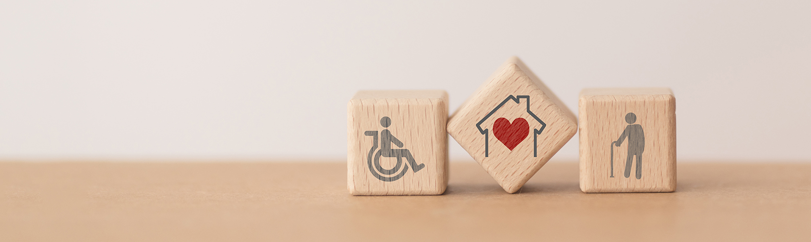 3 Holzblöcke mit Rollstuhlfahrersymbol, rotem Herz in einem Haus und einer Person mit Gehstock